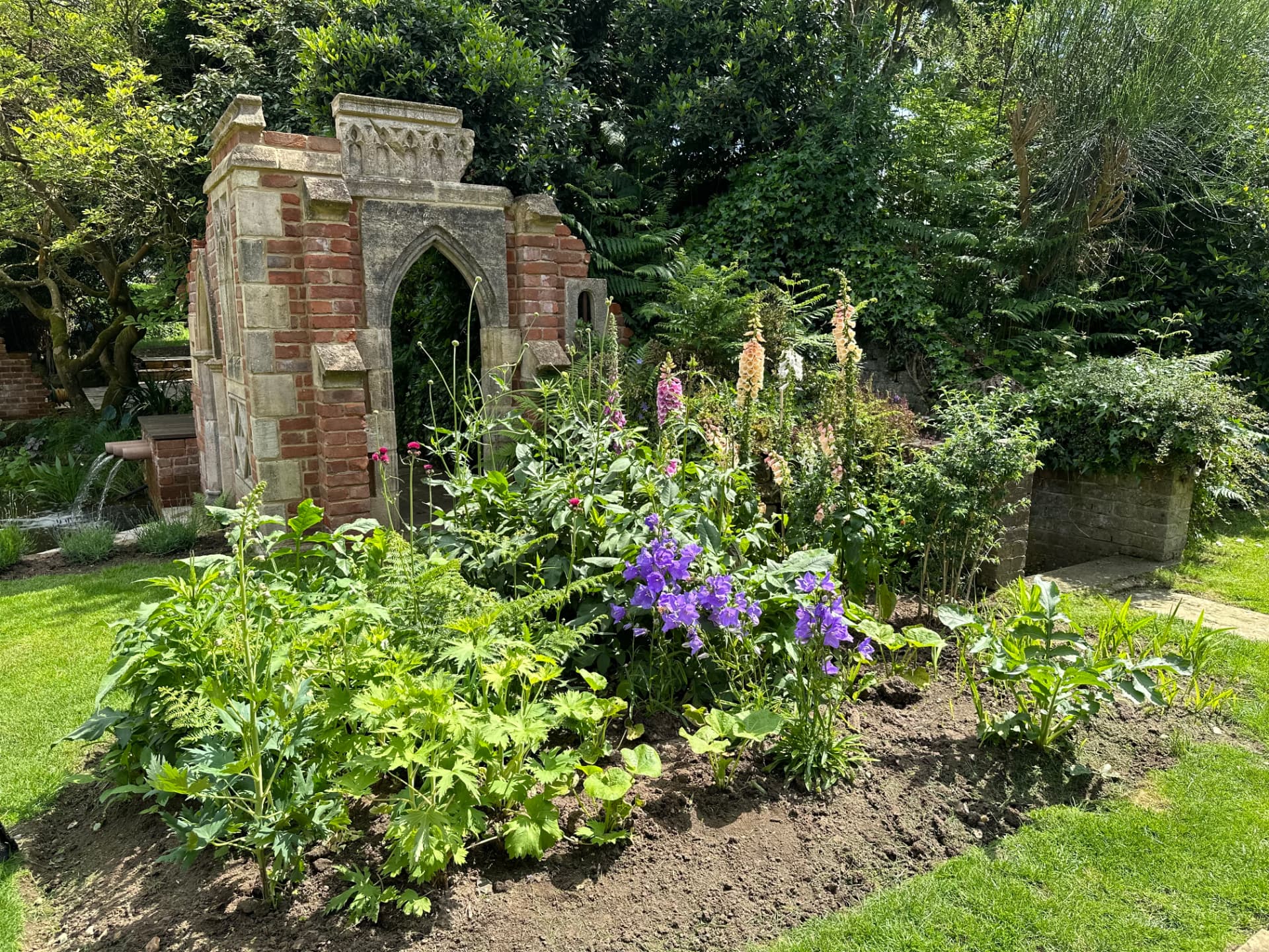 A newly planted cottage-style border with foxgloves and campanula beside a gothic garden folly and lawn
