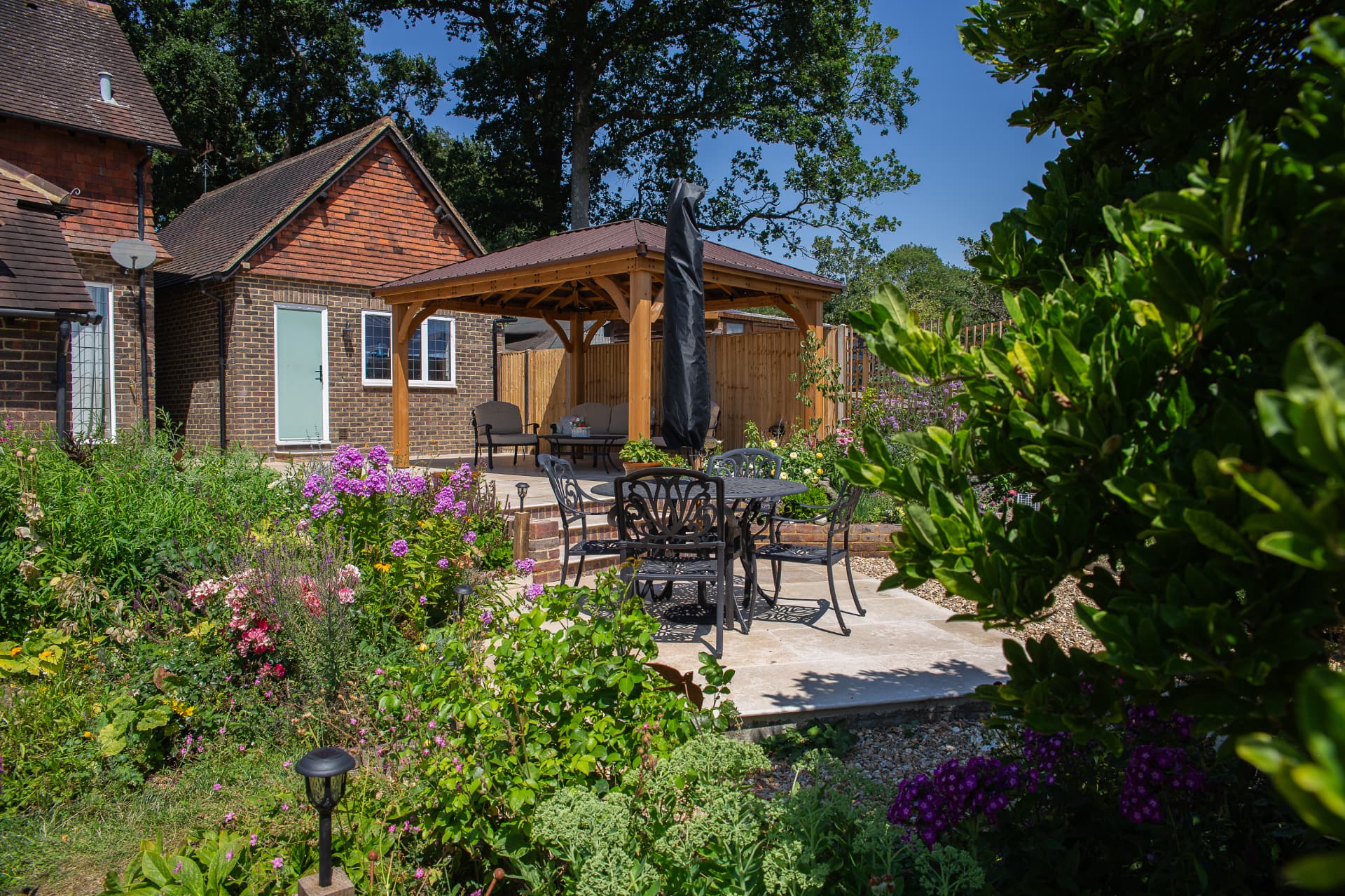 A sunny English garden with richly planted flower borders leading to a stone patio and oak gazebo